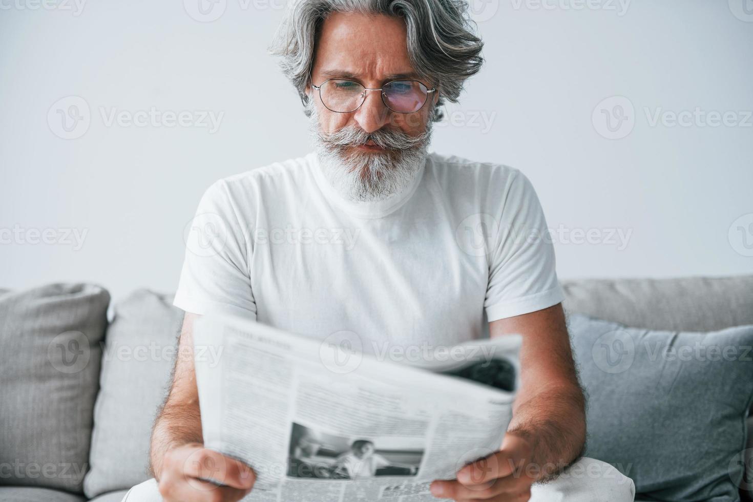 si siede su il divano. anziano elegante moderno uomo con grigio capelli e barba in casa foto