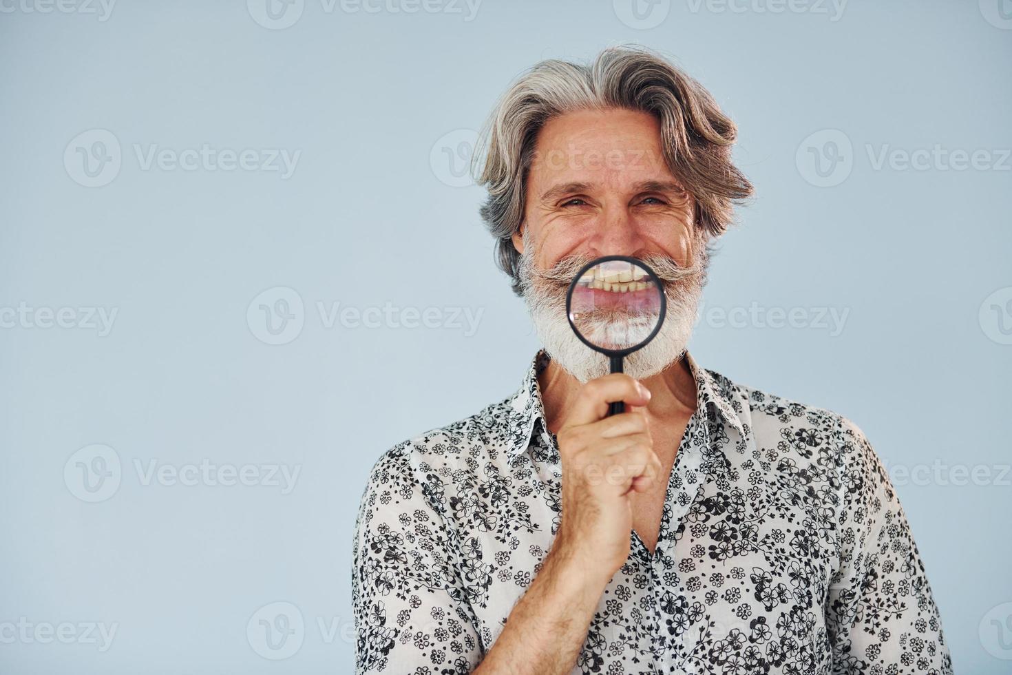 avendo divertimento con ingrandimento bicchiere. anziano elegante moderno uomo con grigio capelli e barba in casa foto