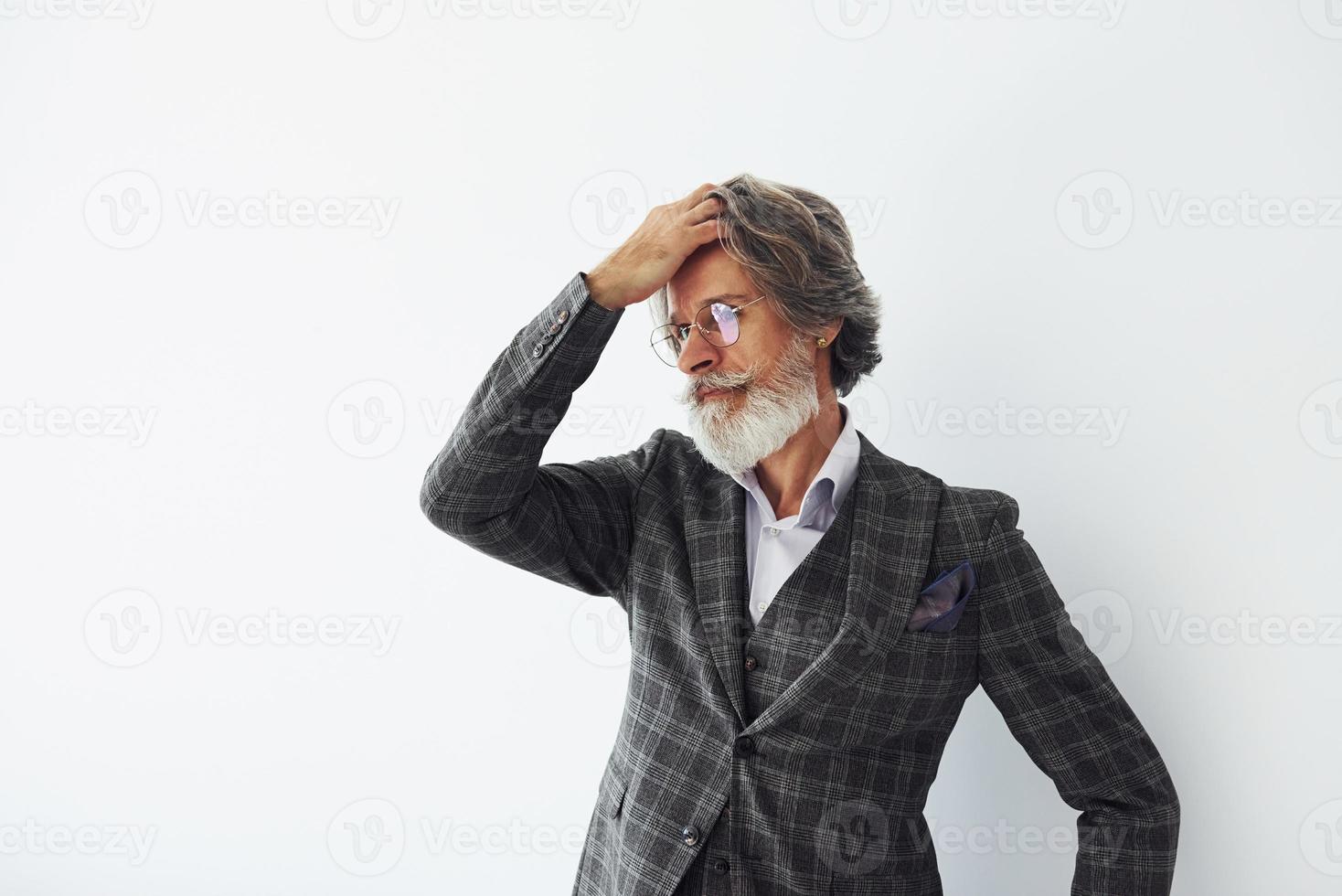 in posa per un' telecamera mentre in piedi contro bianca sfondo. anziano elegante moderno uomo con grigio capelli e barba in casa foto