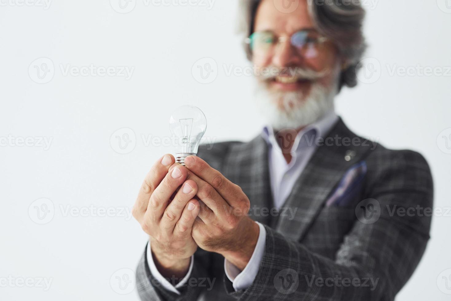 in posa per un' telecamera mentre in piedi contro bianca sfondo. anziano elegante moderno uomo con grigio capelli e barba in casa foto