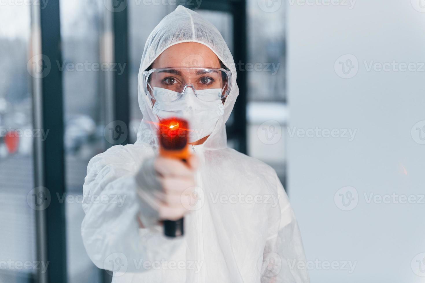 detiene infrarosso termometro. ritratto di femmina medico scienziato nel laboratorio cappotto, difensiva occhiali e maschera foto
