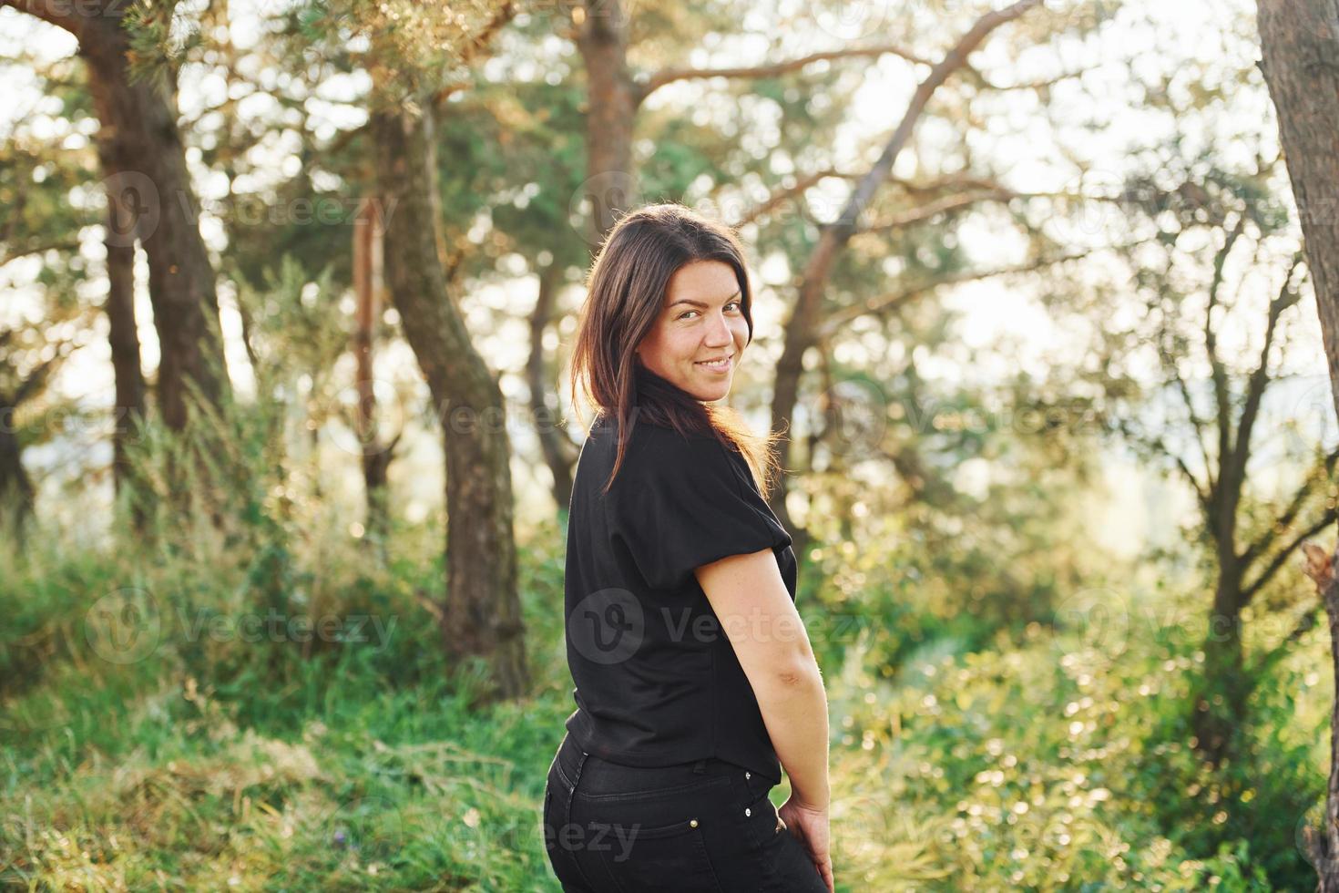 brunetta nel nero camicia e jeans in piedi e in posa nel il foresta a giorno con bellissimo luce del sole foto