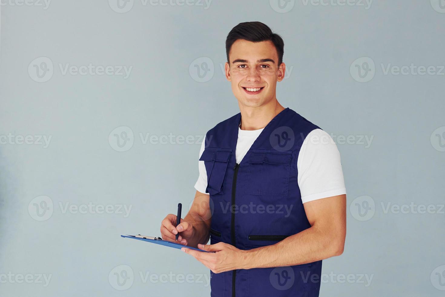 scrive nel bloc notes. maschio lavoratore nel blu uniforme in piedi dentro di studio contro bianca sfondo foto