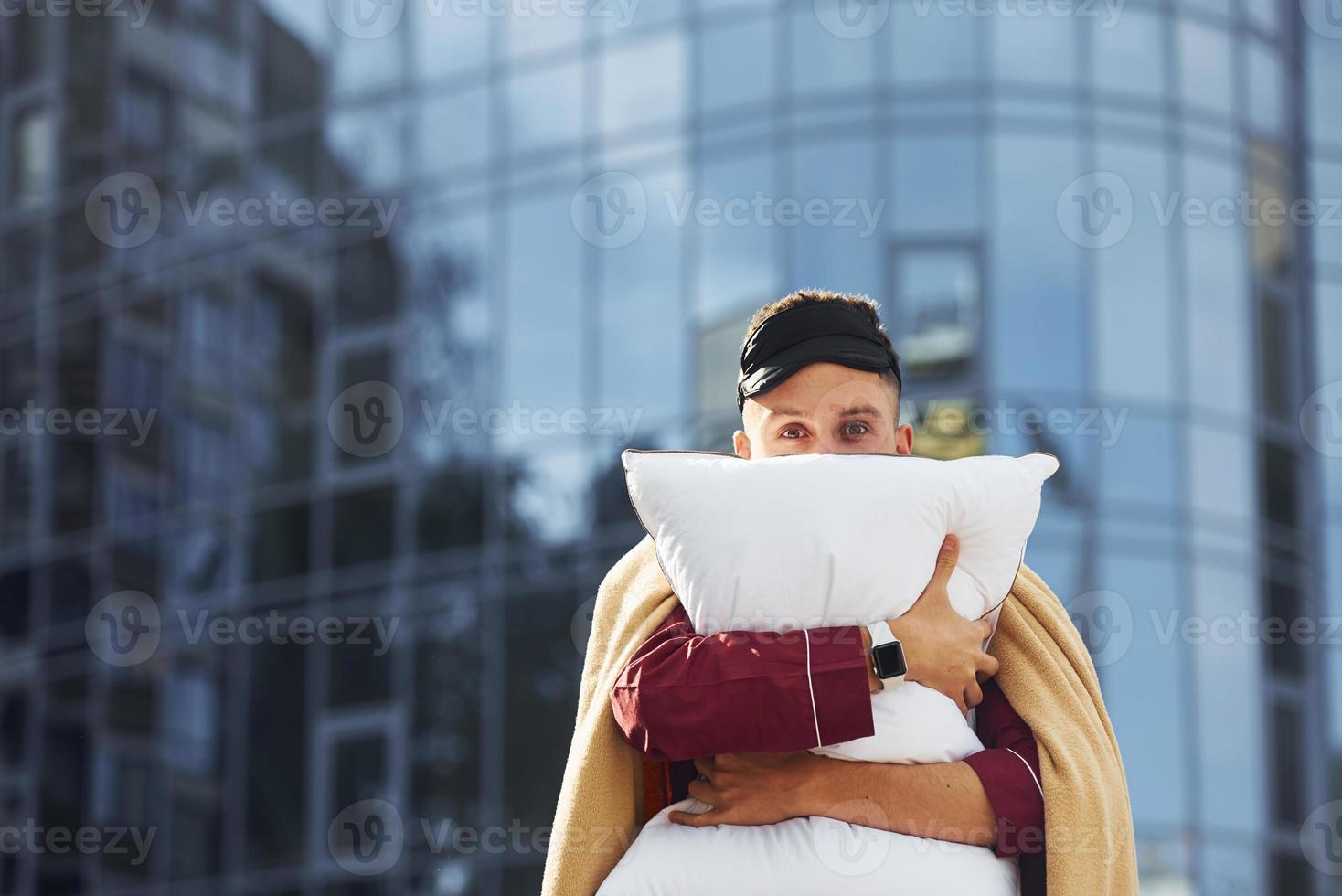 Abbracciare una persona il cuscino. giovane uomo nel pigiama è all'aperto su il strada. si sente assonnato foto