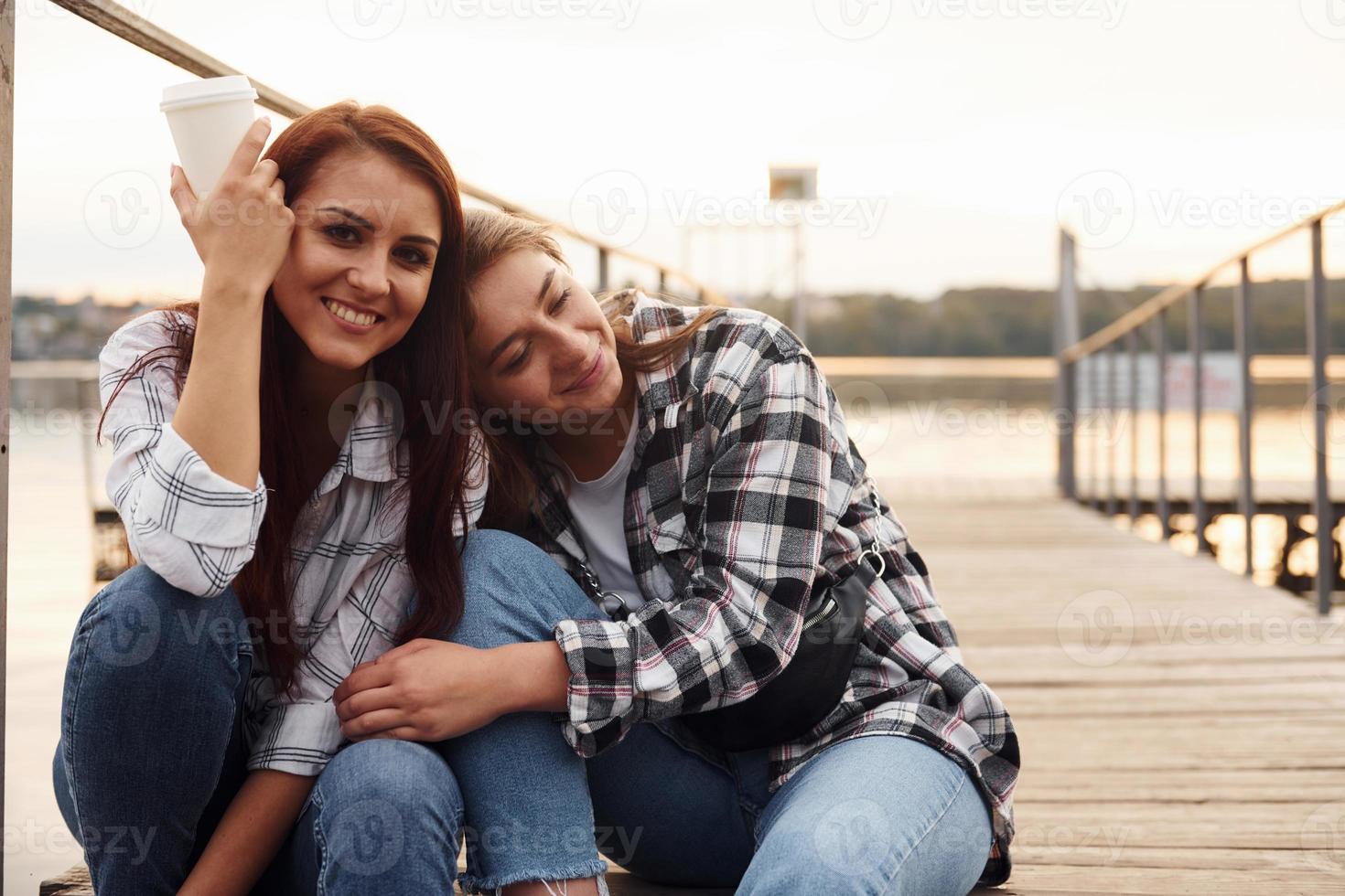 carino lesbica coppia si siede insieme vicino il lago con tazze di bevanda nel mani e Abbracciare una persona ogni altro foto