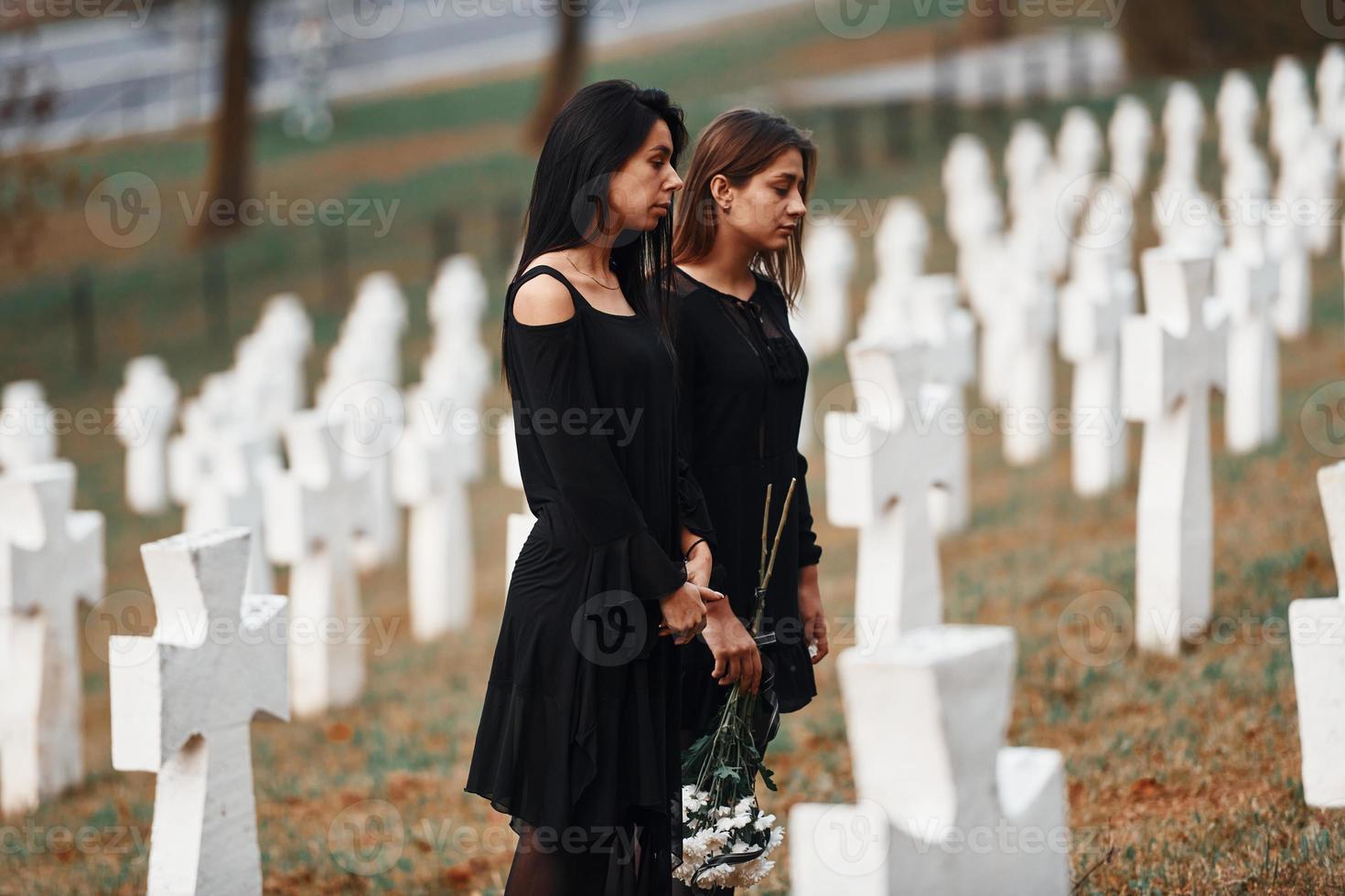 detiene fiori. Due giovane donne nel nero Abiti visitare cimitero con molti bianca croci. concezione di funerale e Morte foto