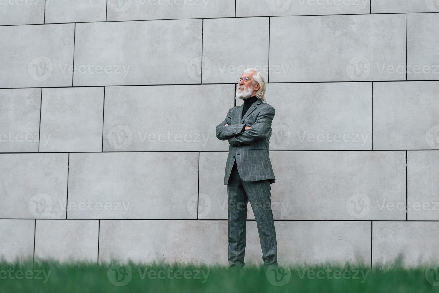 anziano uomo d'affari nel formale Abiti, con grigio capelli e barba è all'aperto sta contro il parete foto