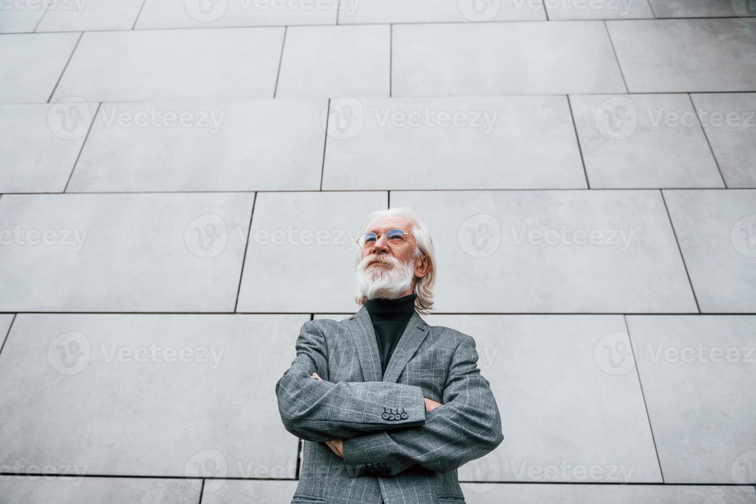 anziano uomo d'affari nel formale Abiti, con grigio capelli e barba è all'aperto sta contro il parete foto