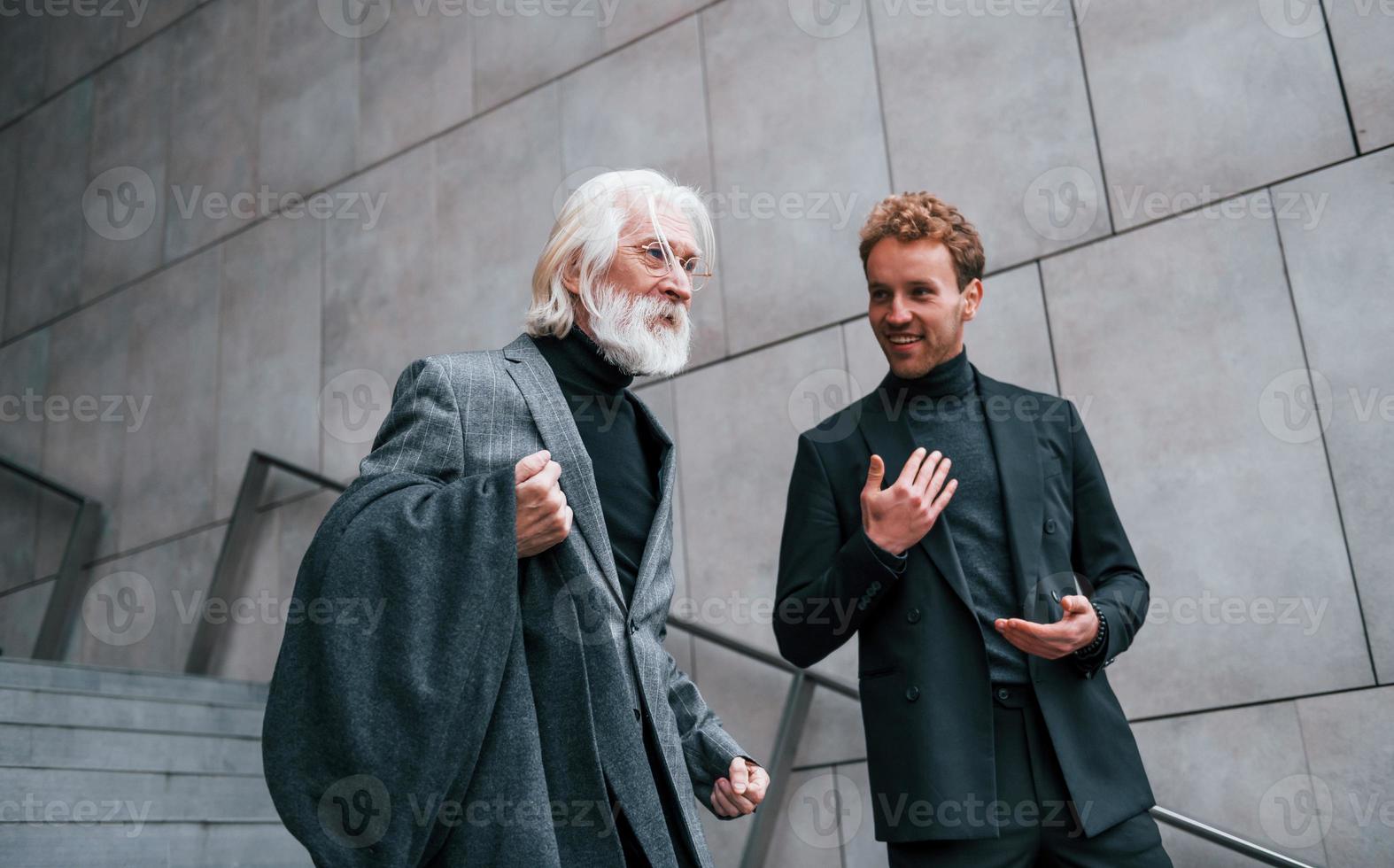 a piedi su il scale. giovane tipo con anziano uomo nel elegante Abiti è all'aperto insieme. concezione di attività commerciale foto
