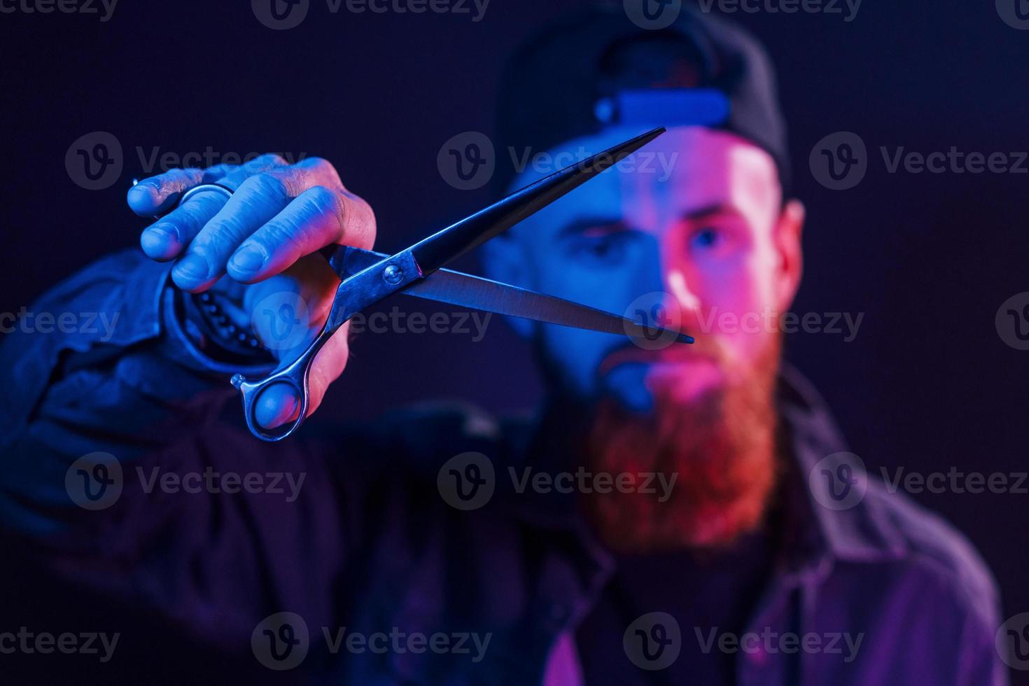 con forbici. giovane barbuto barbiere nel berretto in piedi nel il studio con neon illuminazione foto