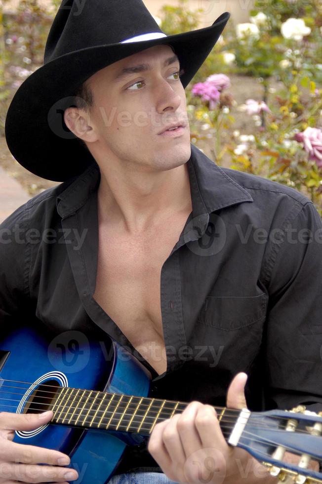 un' bello cowboy canta con il suo blu chitarra nel un' parco pieno di Rose. foto