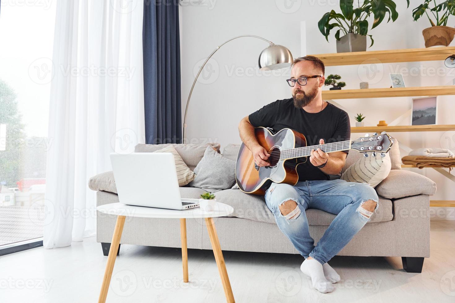 utilizzando il computer portatile. uomo nel casuale Abiti e con acustico chitarra è in casa foto