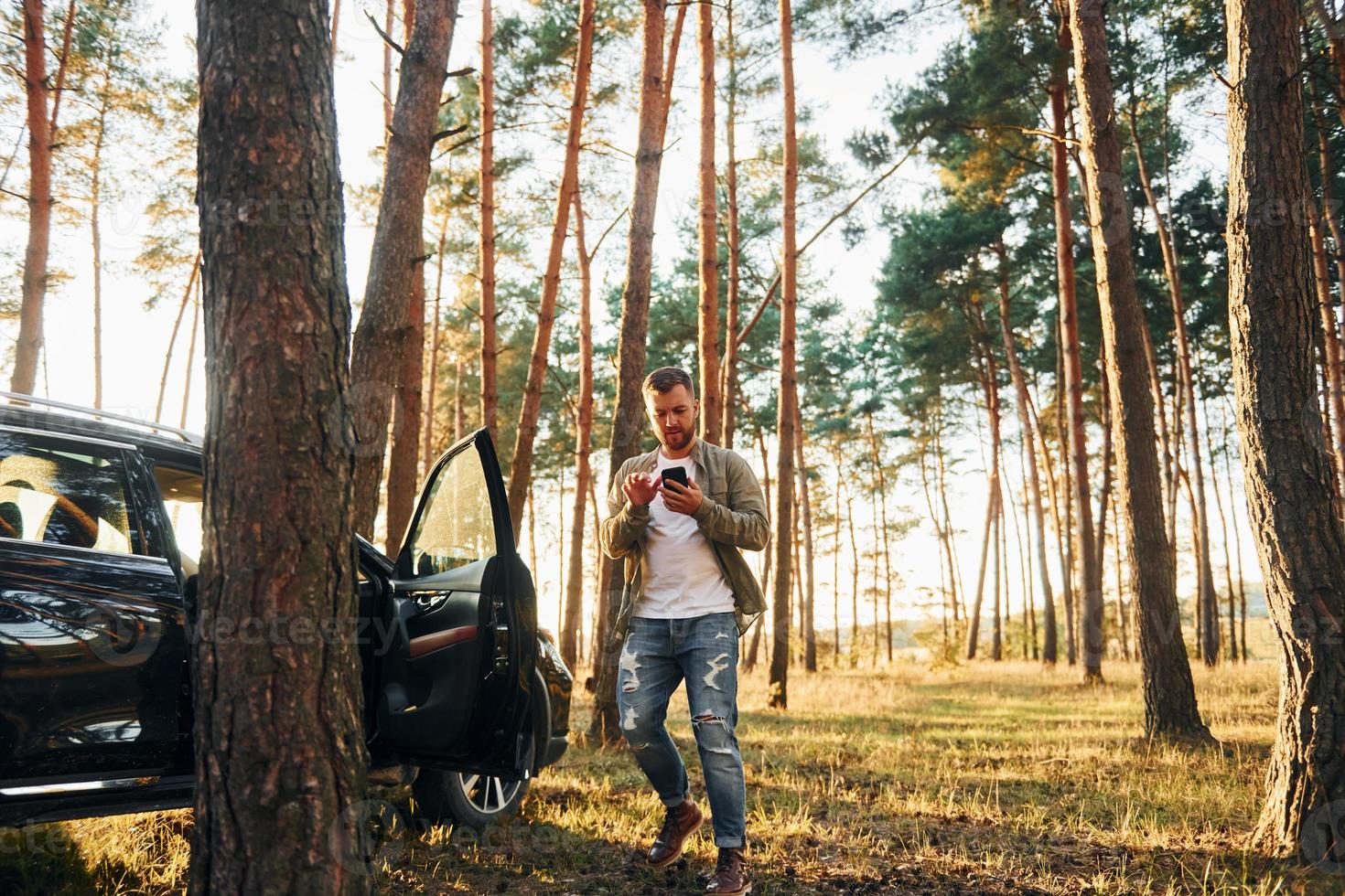 vicino il macchina. uomo nel jeans è all'aperto nel il foresta con il suo nero colorato automobile foto