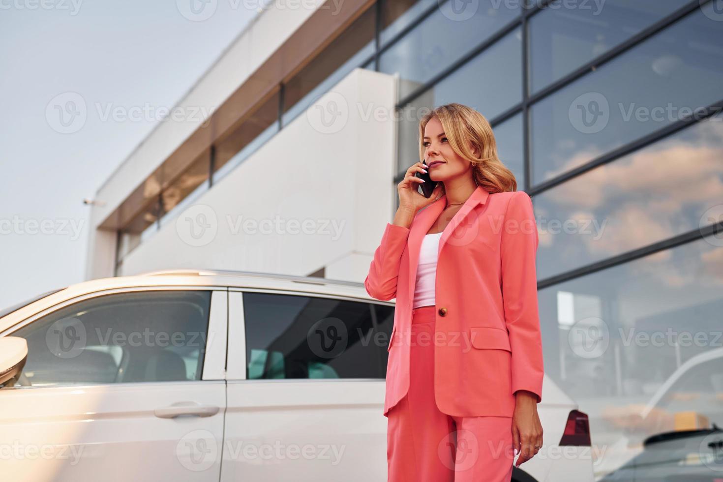 avendo conversazione di Telefono. donna nel formale indossare in piedi all'aperto con Telefono nel mani e vicino auto foto
