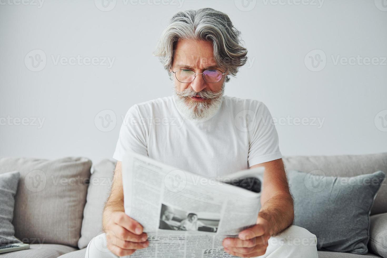 si siede su il divano. anziano elegante moderno uomo con grigio capelli e barba in casa foto