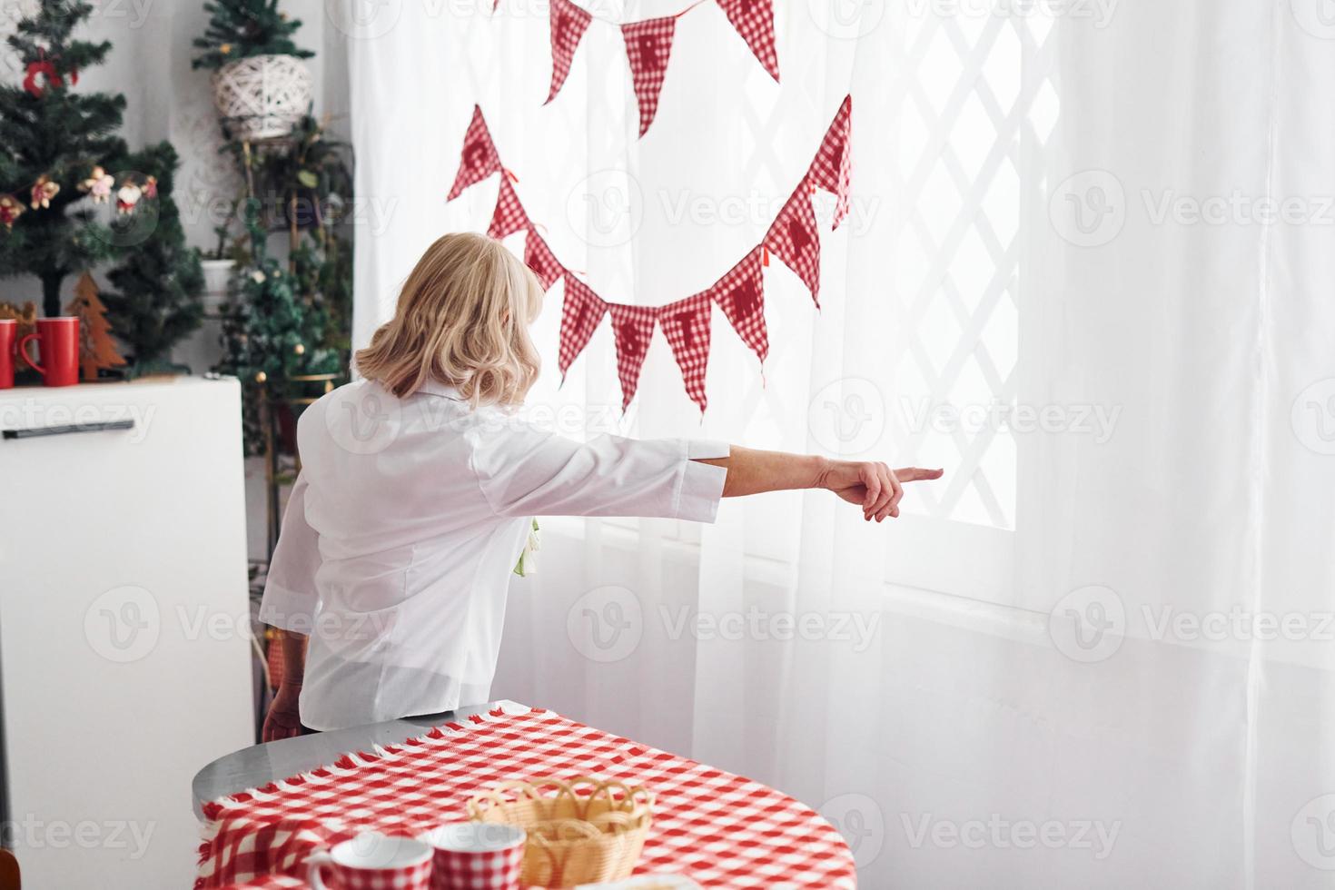 posteriore Visualizza di anziano donna nel bianca Abiti quello in piedi su il cucina e puntamento per il giusto di mano foto