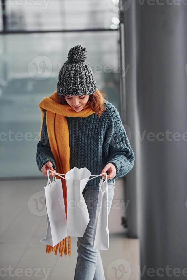 bellissimo allegro ragazza nel giallo sciarpa e nel caldo Abiti in piedi in casa con shopping borse nel mani foto