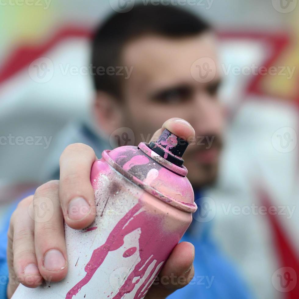 un' giovane graffiti artista nel un' blu giacca è Tenere un' può di dipingere nel davanti di lui contro un' sfondo di colorato graffiti disegno. strada arte e vandalismo concetto foto