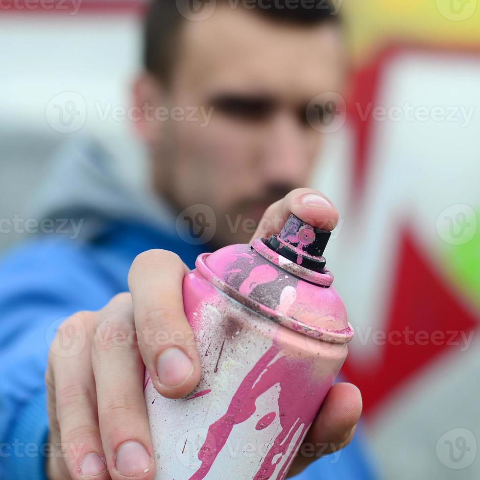 un' giovane graffiti artista nel un' blu giacca è Tenere un' può di dipingere nel davanti di lui contro un' sfondo di colorato graffiti disegno. strada arte e vandalismo concetto foto