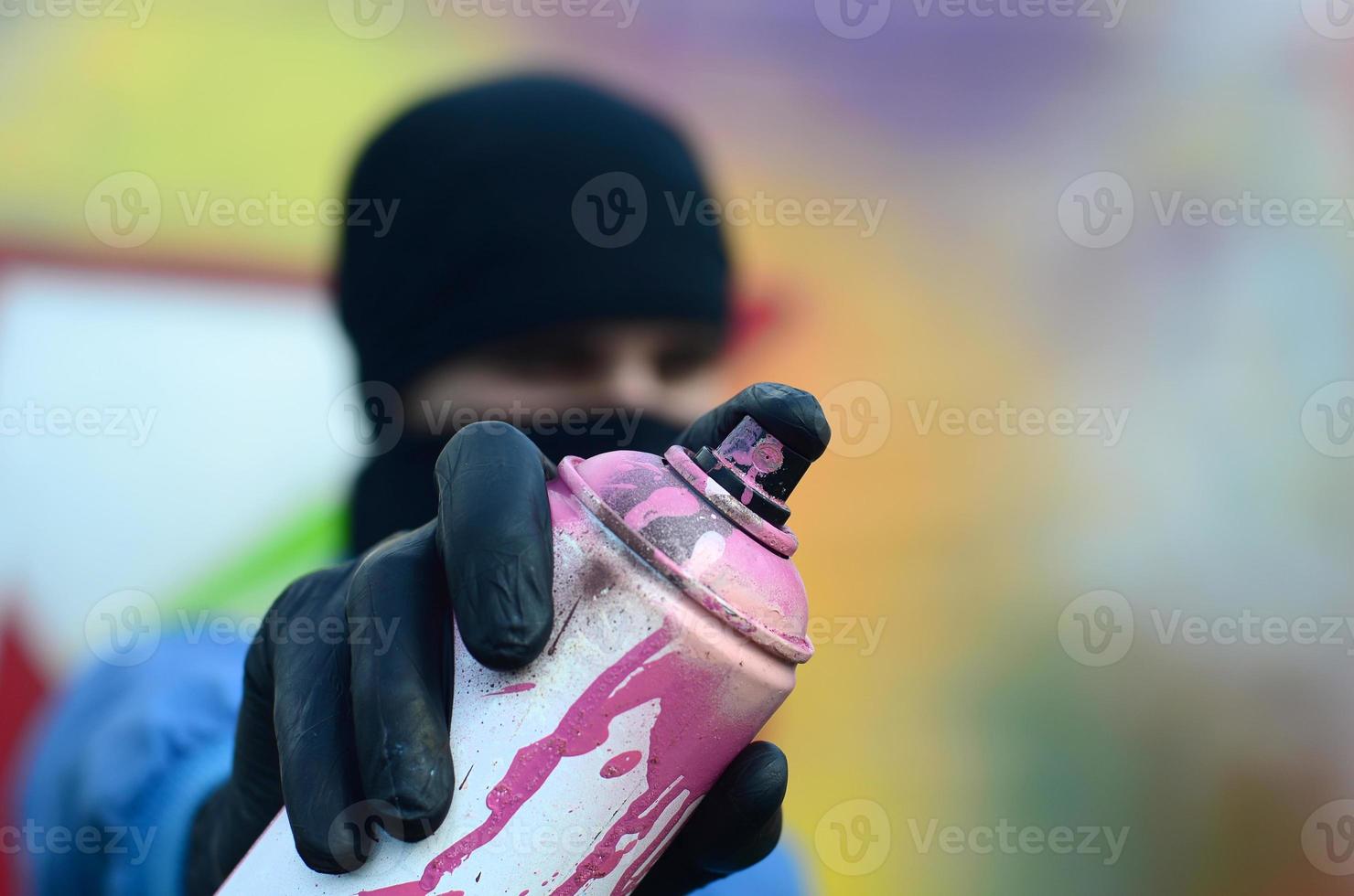 un' giovane graffiti artista nel un' blu giacca e nero maschera è Tenere un' può di dipingere nel davanti di lui contro un' sfondo di colorato graffiti disegno. strada arte e vandalismo concetto foto