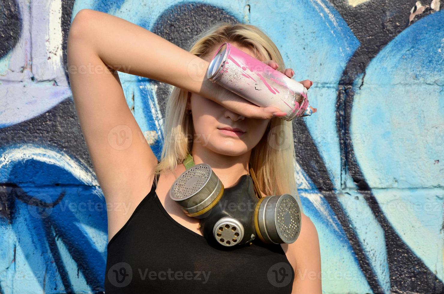 giovane e bellissimo sexy ragazza graffiti artista con gas maschera su sua collo nascondiglio il suo occhi con un' spray può in piedi su un' parete sfondo con un' graffiti modello nel blu e viola toni foto