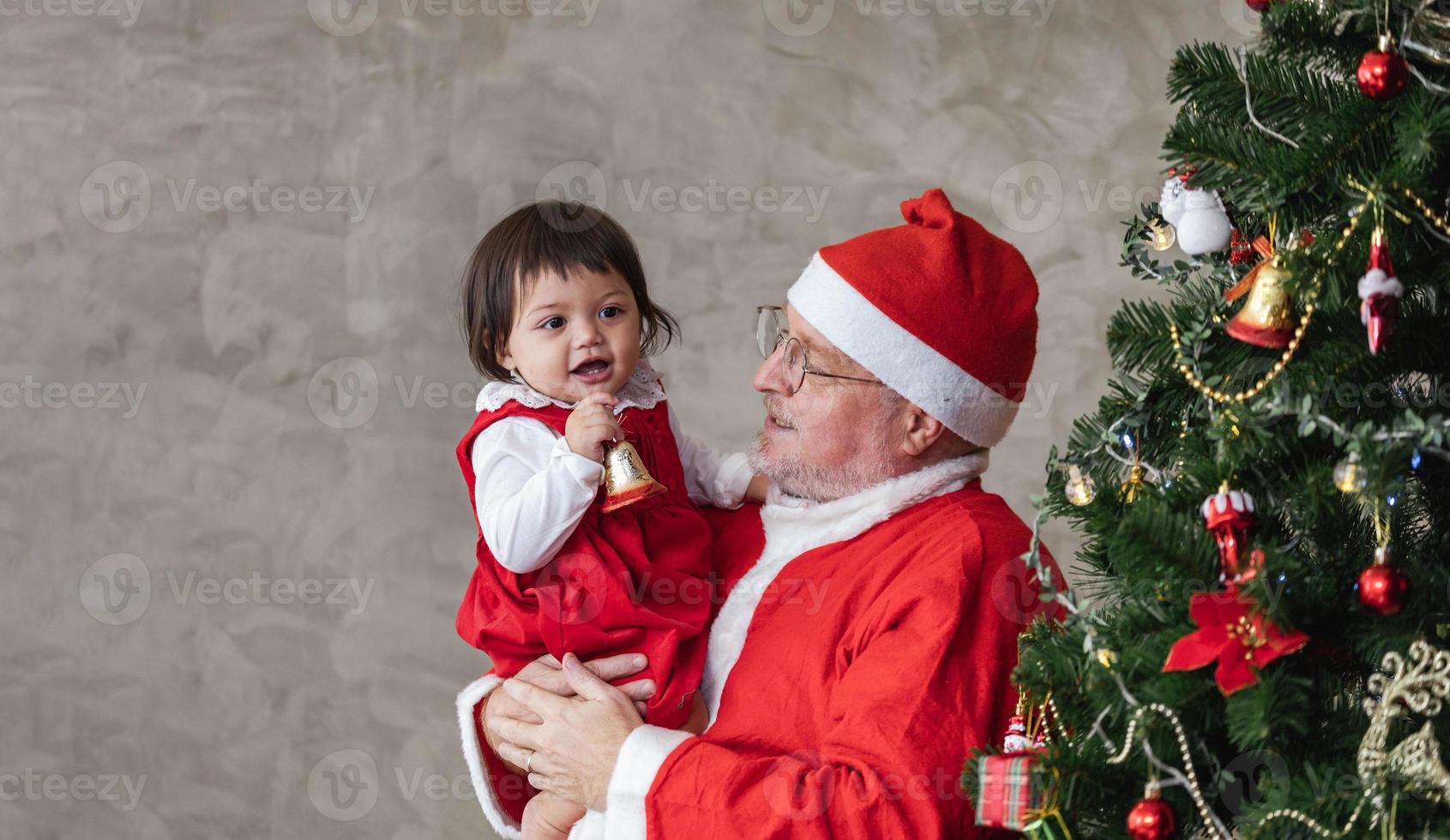 Santa Claus è sollevamento contento poco bambino piccolo bambino ragazza su e ridendo allegramente mentre porzione per decorare Natale albero su il indietro per stagione celebrazione foto