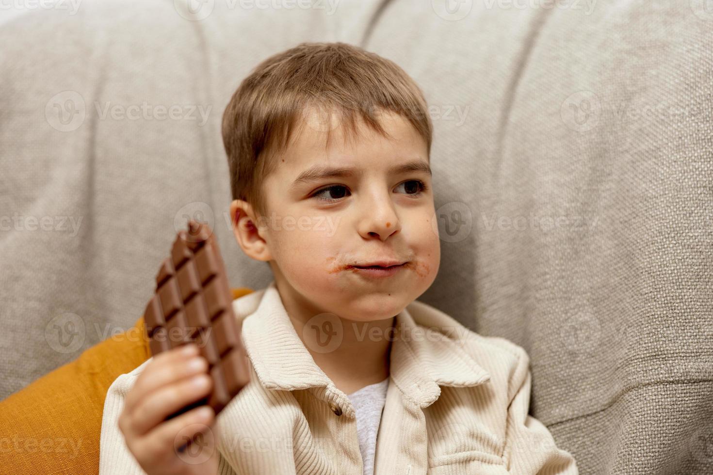 poco adorabile ragazzo seduta su il divano a casa e mangiare cioccolato sbarra. bambino e dolci, zucchero confetteria. ragazzo godere un' delizioso dolce. prescolastico bambino con casuale vestiario. positivo emozione. foto