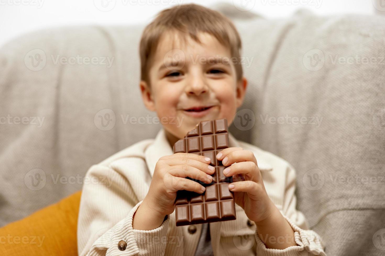 poco adorabile ragazzo seduta su il divano a casa e mangiare cioccolato sbarra. bambino e dolci, zucchero confetteria. ragazzo godere un' delizioso dolce. prescolastico bambino con casuale vestiario. positivo emozione. foto