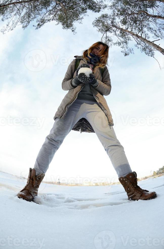 un' giovane e gioioso caucasico ragazza nel un' Marrone cappotto detiene un' palla di neve nel davanti di un' orizzonte linea fra il cielo e un' congelato lago nel inverno. fisheye foto