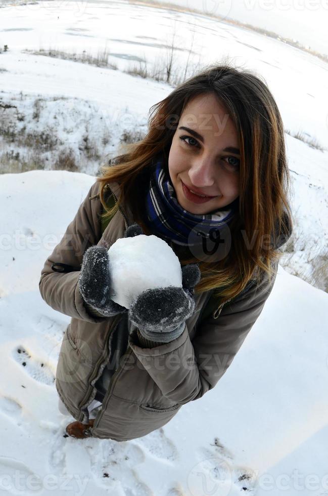 un' giovane e gioioso caucasico ragazza nel un' Marrone cappotto detiene un' palla di neve nel davanti di un' orizzonte linea fra il cielo e un' congelato lago nel inverno. fisheye foto