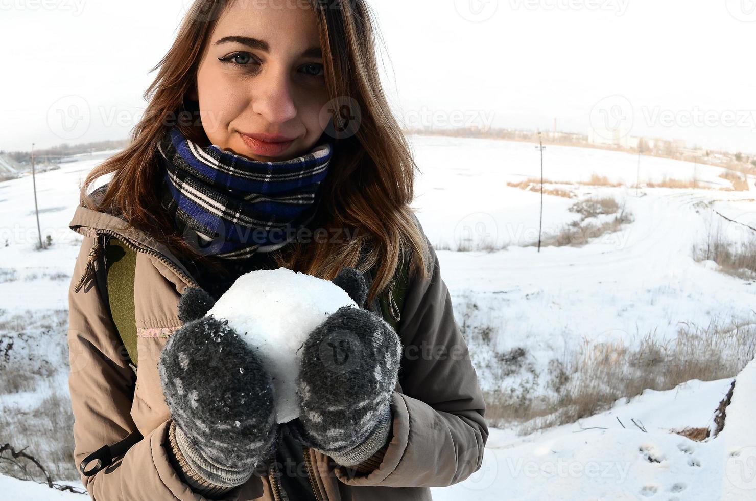 un' giovane e gioioso caucasico ragazza nel un' Marrone cappotto detiene un' palla di neve nel davanti di un' orizzonte linea fra il cielo e un' congelato lago nel inverno. fisheye foto