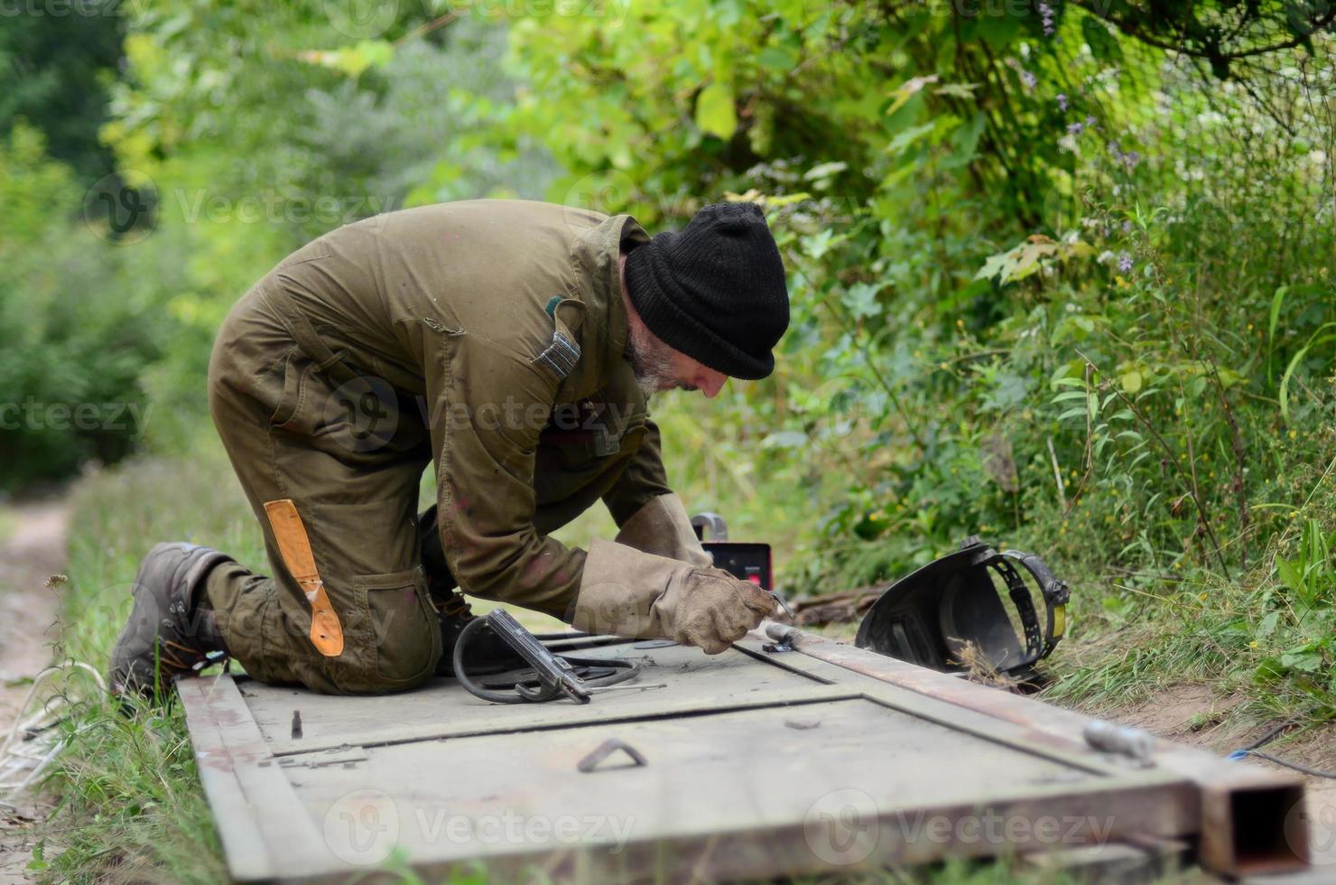 vecchio uomo saldatore nel Marrone uniforme prepara metallo porta superficie per saldatura con arco saldatura macchina all'aperto foto