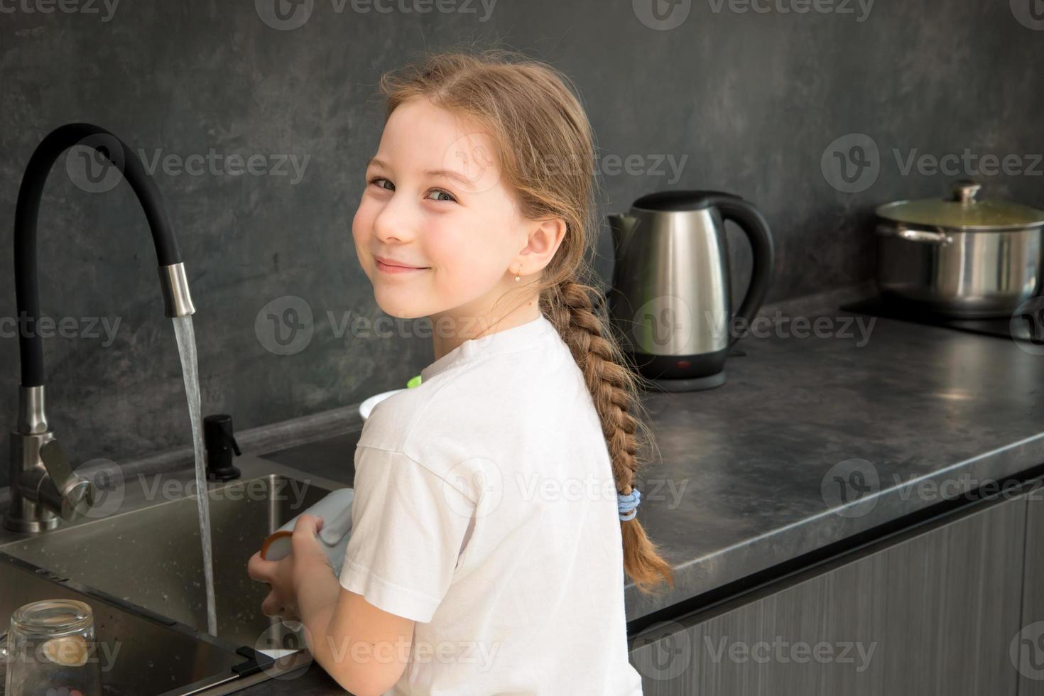 carino poco ragazza con un' treccia lavaggi piatti nel il cucina a il Lavello e sorrisi foto