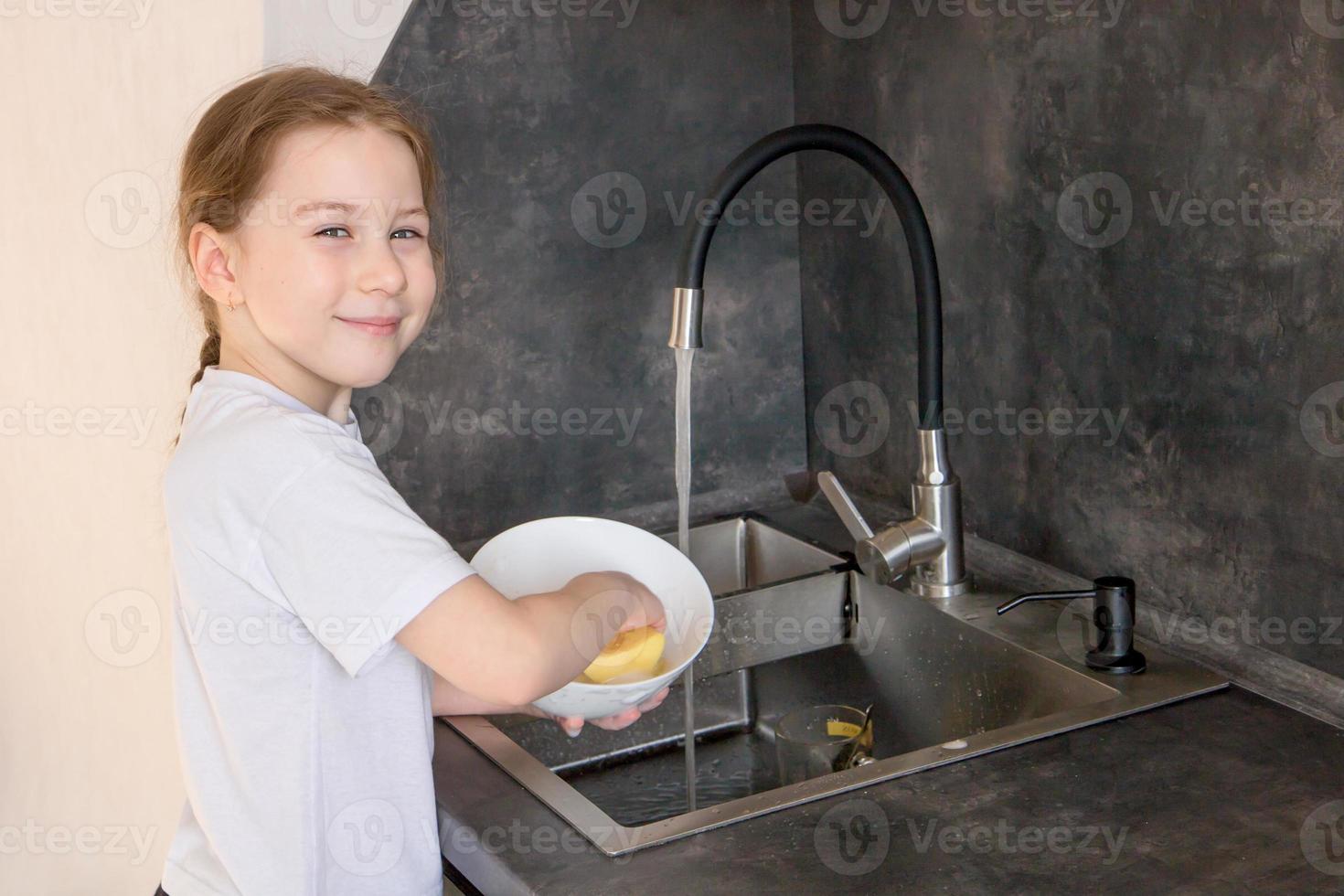 carino poco ragazza con un' treccia lavaggi piatti nel il cucina a il Lavello e sorrisi foto