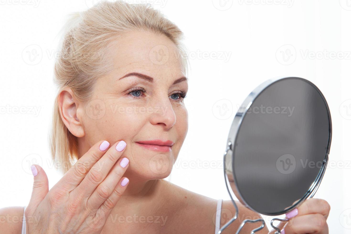 contento mezzo anziano donna modello toccante viso pelle guardare nel specchio. sorridente maturo più vecchio signora godendo salutare pelle cura, invecchiamento bellezza, cura della pelle trattamento cosmetico prodotti concetto. foto