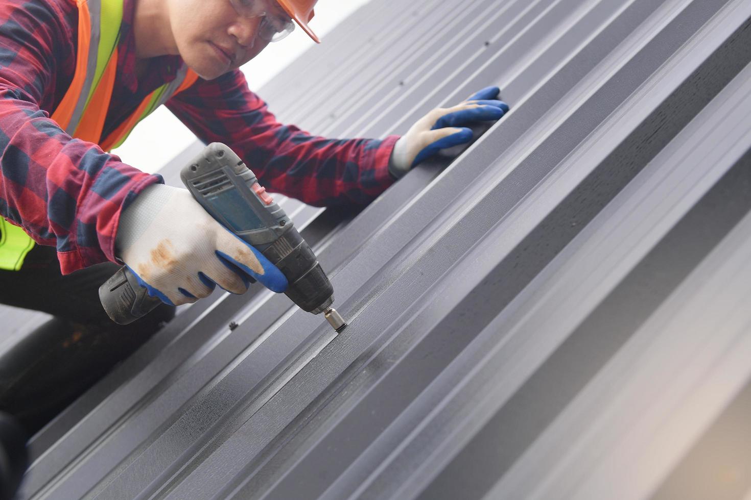 vicino su trapano roofer lavoratore nel protettivo uniforme indossare e guanti, coperture strumenti, installazione nuovo tetti sotto costruzione, elettrico trapano Usato su nuovo tetti con metallo foglio. foto