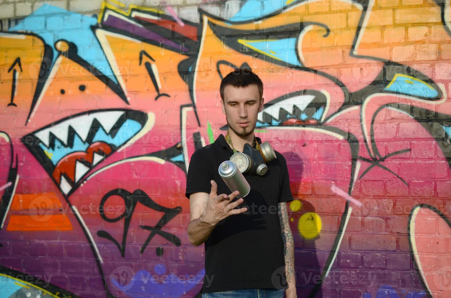 giovane graffiti artista con gas maschera su il suo collo gettare il suo spray può contro colorato rosa graffiti su mattone parete. strada arte e contemporaneo pittura processi foto