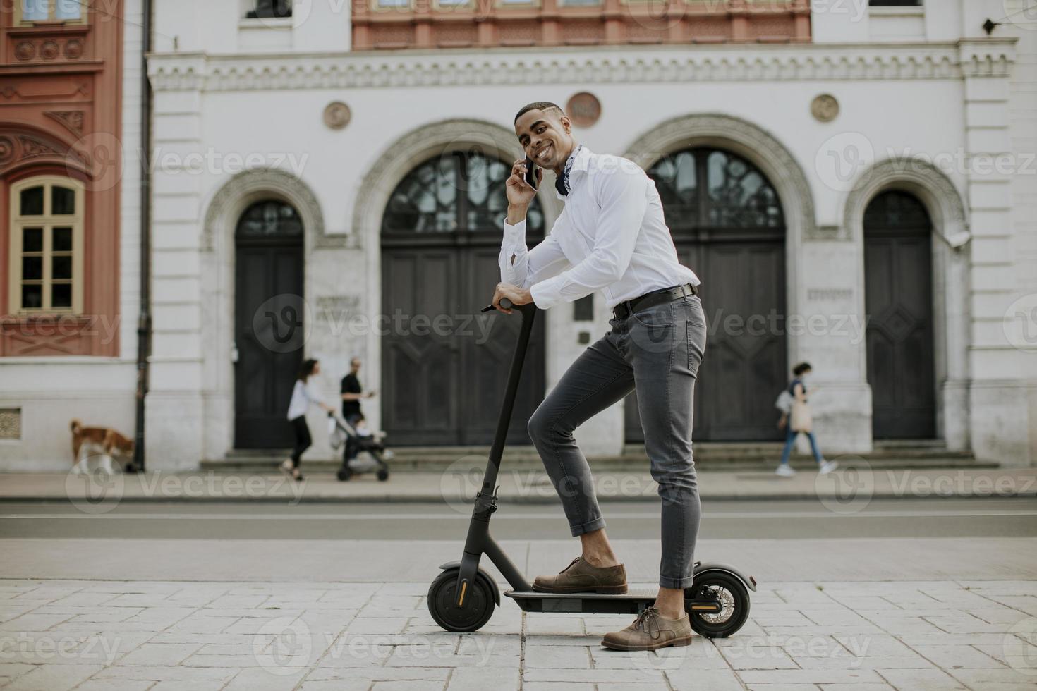 giovane afroamericano che utilizza il telefono cellulare mentre sta in piedi con uno scooter elettrico su una strada foto