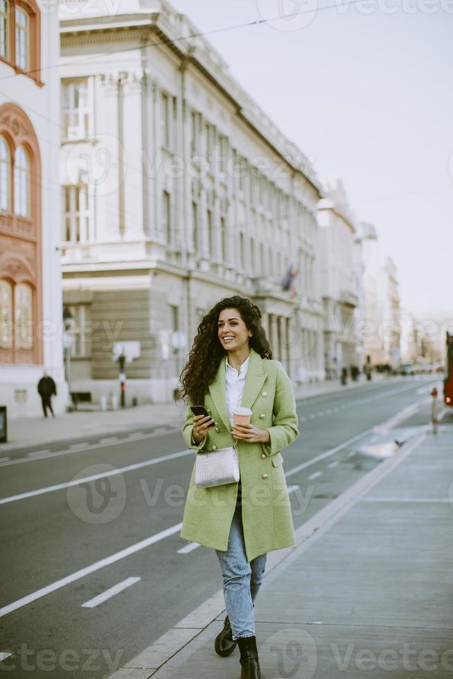 giovane donna che utilizza smartphone per strada e tiene caffè da asporto foto