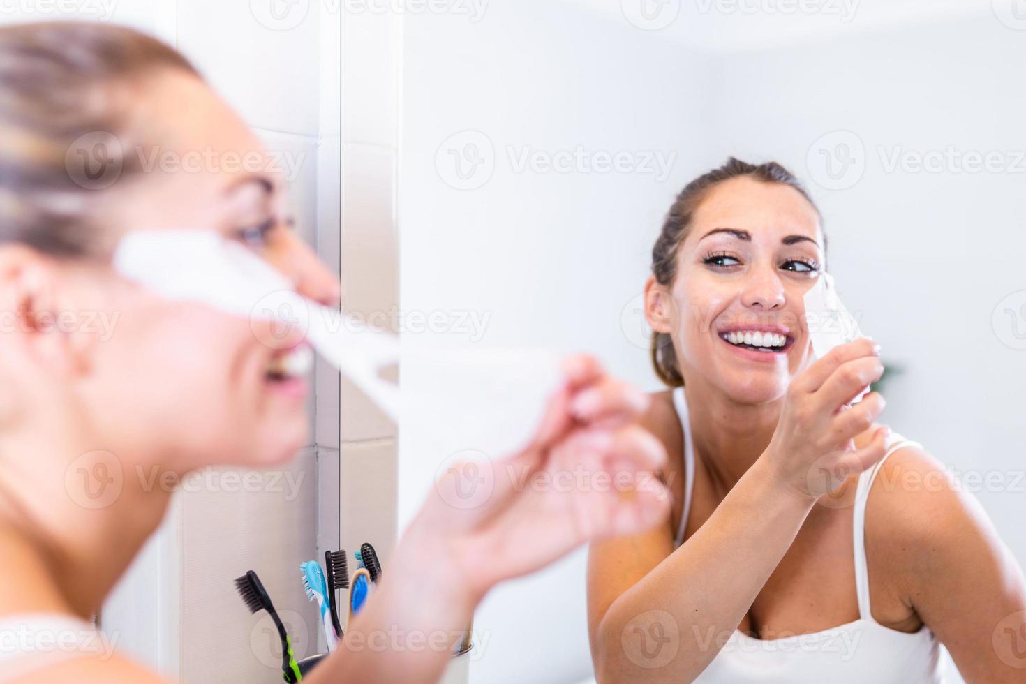 donna sembra nel specchio e assunzione via cosmetico maschera. facciale detergente a casa concetto. pelle cura ragazza toccare cerotti di tessuto maschera sotto occhi per ridurre occhio borse. foto