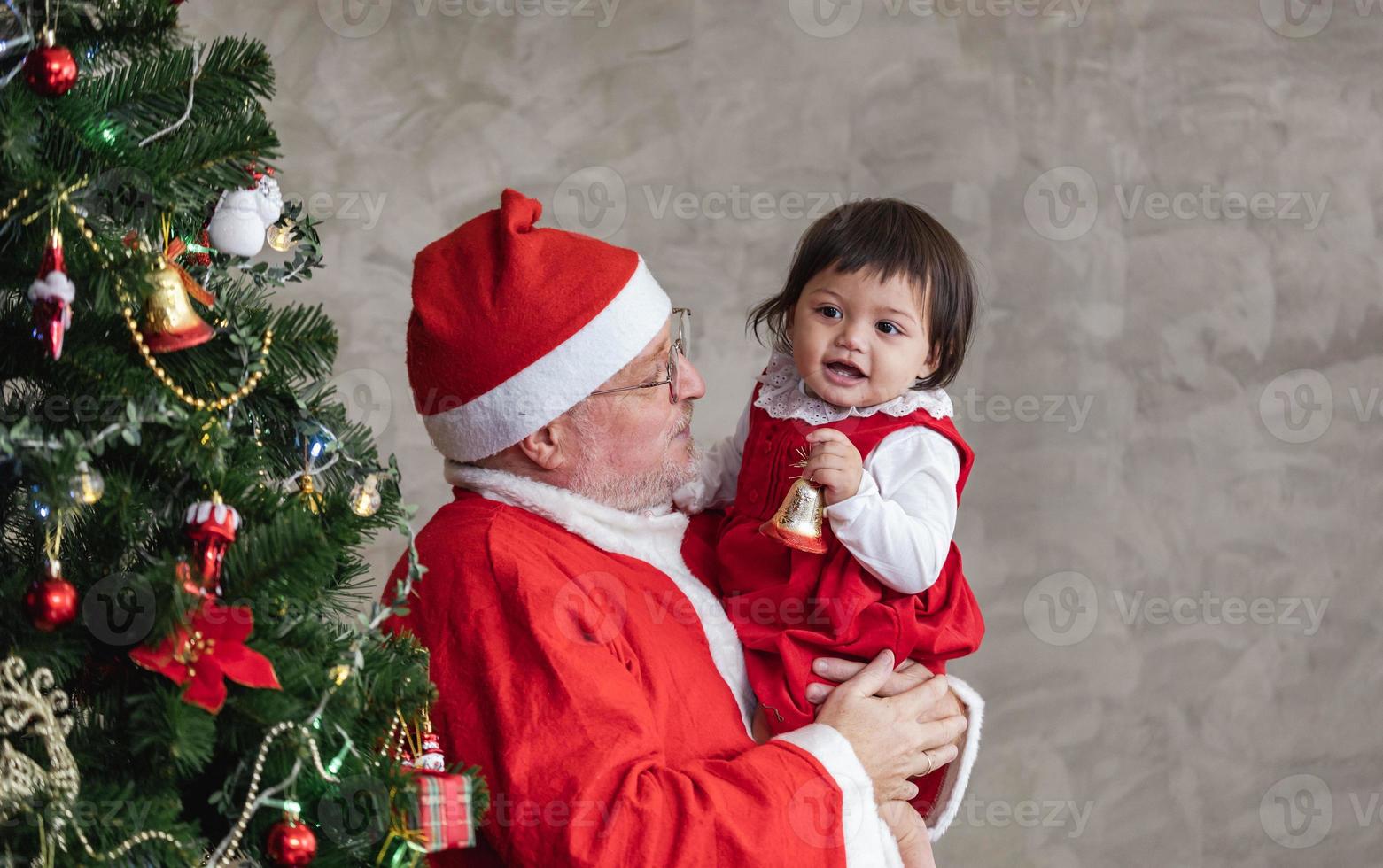 Santa Claus è sollevamento contento bambino piccolo bambino ragazza su e ridendo allegramente mentre porzione per decorare Natale albero su il indietro per stagione celebrazione concetto foto