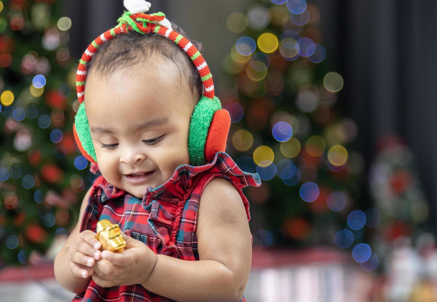 africano americano bambino è felicemente sorridente come giocando con ornamento fronzolo mentre condimento nel Natale vestito e Santa cappello con Natale albero su il indietro per stagione celebrazione concetto foto