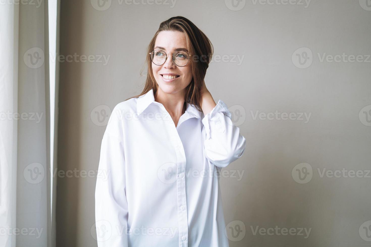 ritratto di giovane sorridente brunetta donna nel occhiali e bianca camicia vicino grigio parete foto