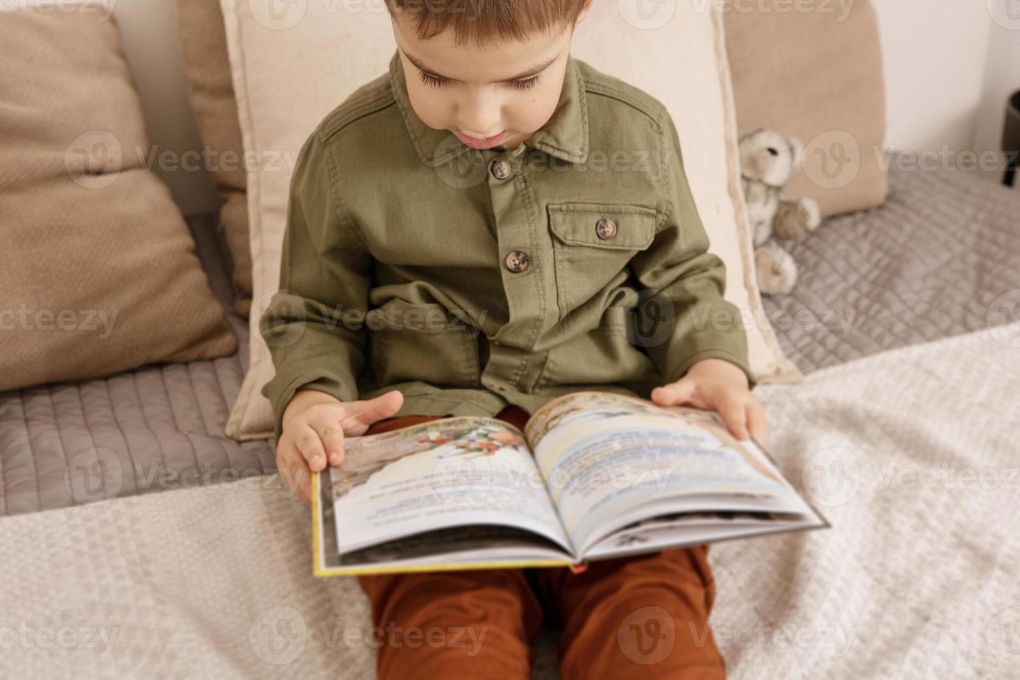 poco e carino caucasico ragazzo lettura un' libro su il letto a casa. interno e Abiti nel naturale terra colori. accogliente ambiente. bambino legge un' Fata racconto. selettivo messa a fuoco. foto