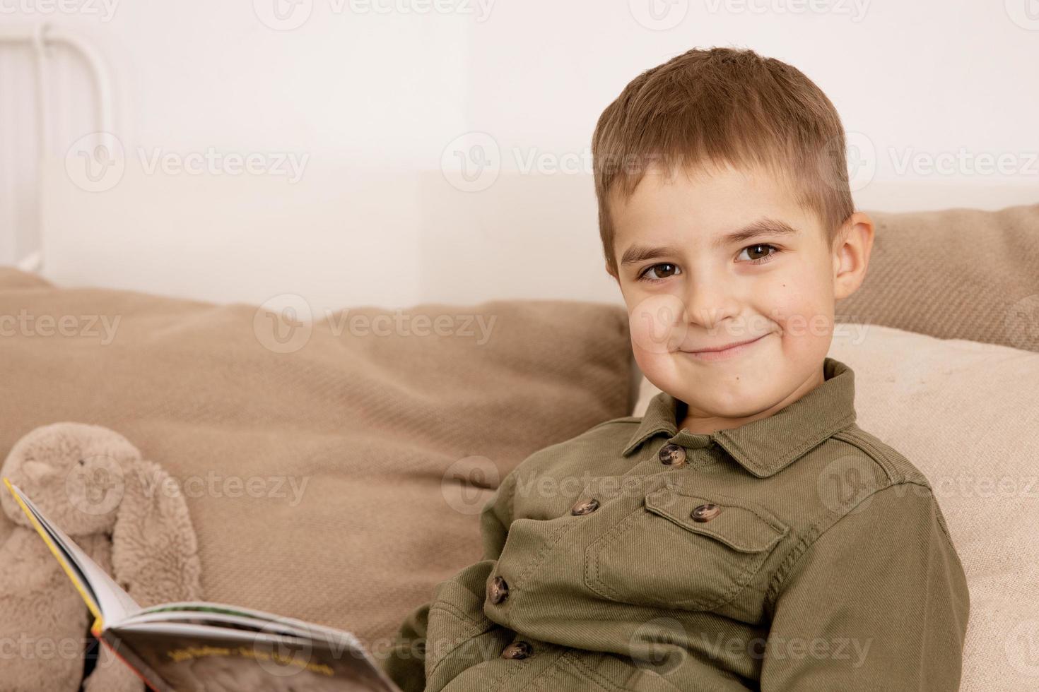 poco e carino caucasico ragazzo lettura un' libro su il letto a casa. interno e Abiti nel naturale terra colori. accogliente ambiente. bambino legge un' Fata racconto. foto