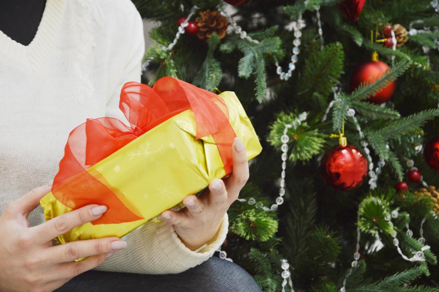 il ragazza detiene nel sua mani un' regalo scatola nel un' d'oro pacchetto con un' rosso arco su il sfondo di un' decorato Natale albero. vacanza. regalo avvolgere. foto