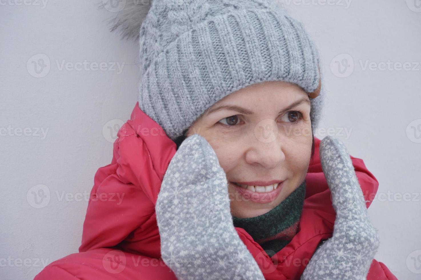 un' giovane donna nel un' a maglia grigio cappello e guanti sorrisi e sembra in il distanza. ritratto di un' donna nel un' rosso giacca con un' cappuccio. foto