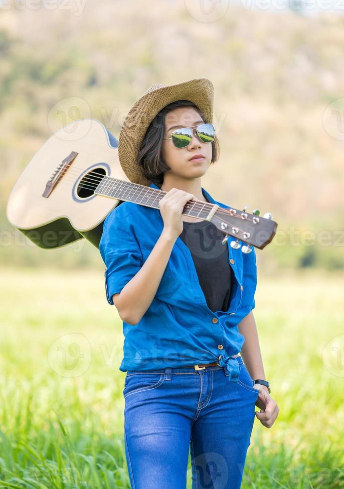 donna indossare cappello e trasportare sua chitarra nel erba campo foto