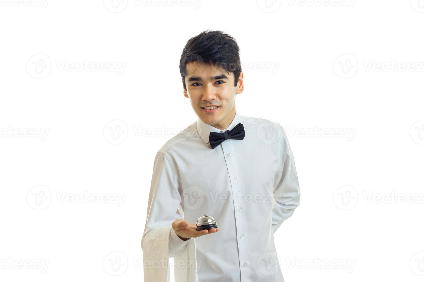 giovane affascinante Cameriere nel un' bianca camicia e nero arco cravatta sembra inoltrare e pendente detiene nel il suo mano campana foto
