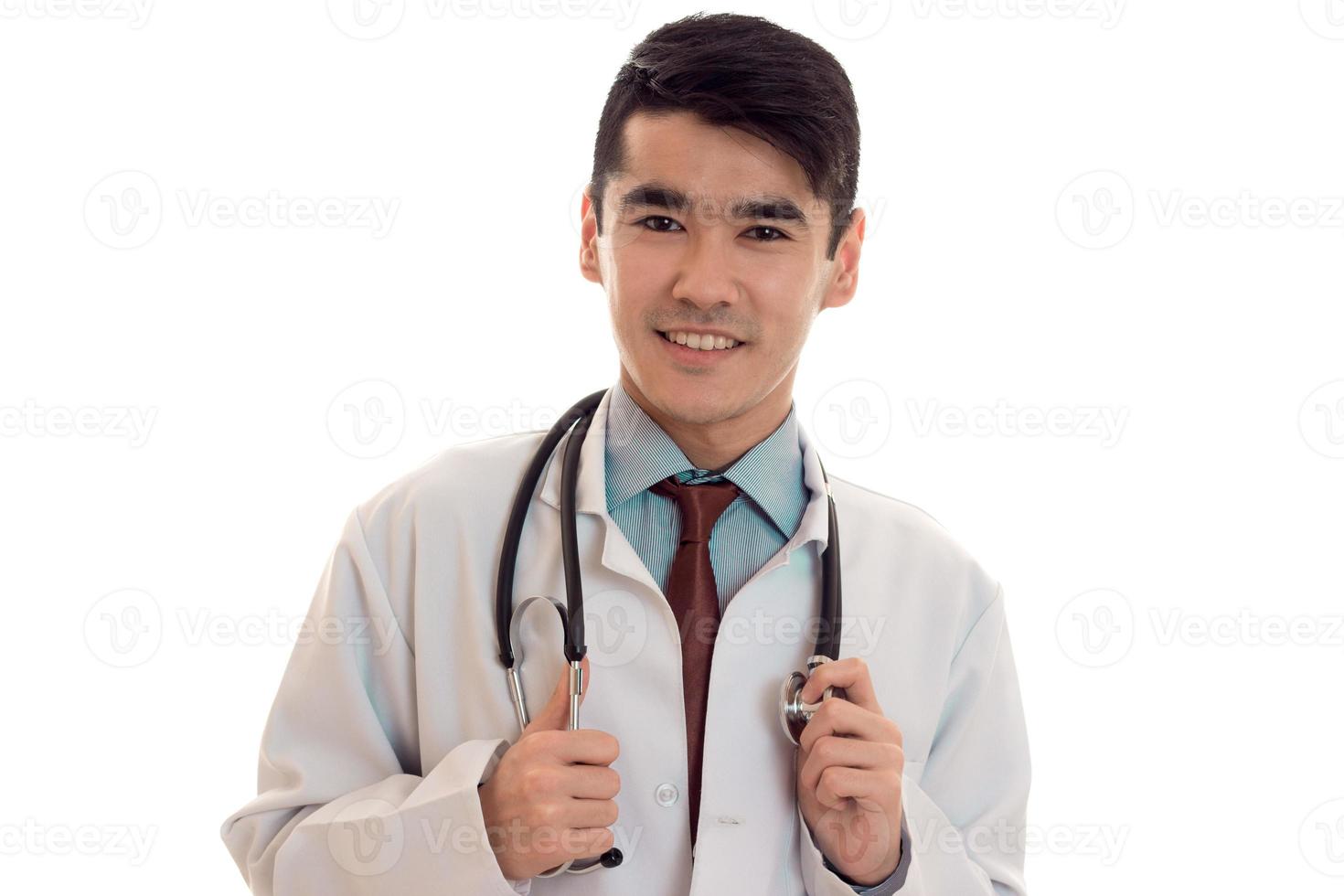ritratto di giovane contento brunetta uomo medico nel bianca uniforme sorridente su telecamera con stetoscopio su il suo collo isolato su vuoto sfondo foto